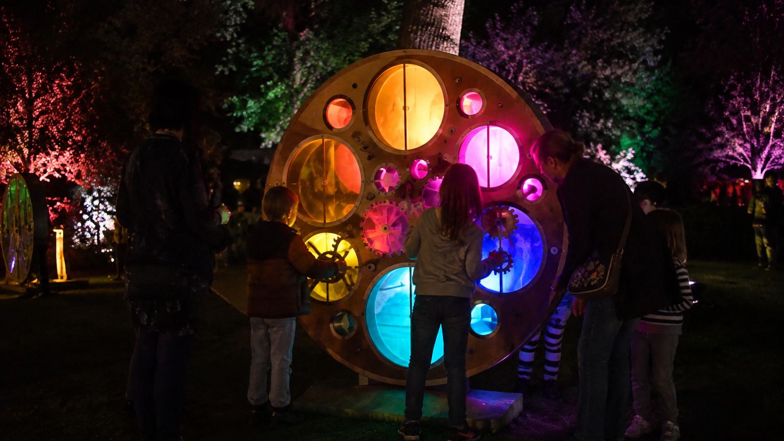 Children and adults gathered around a large illuminated disc of coloured glass and turning cogs in a garden at night, several of them working it together.