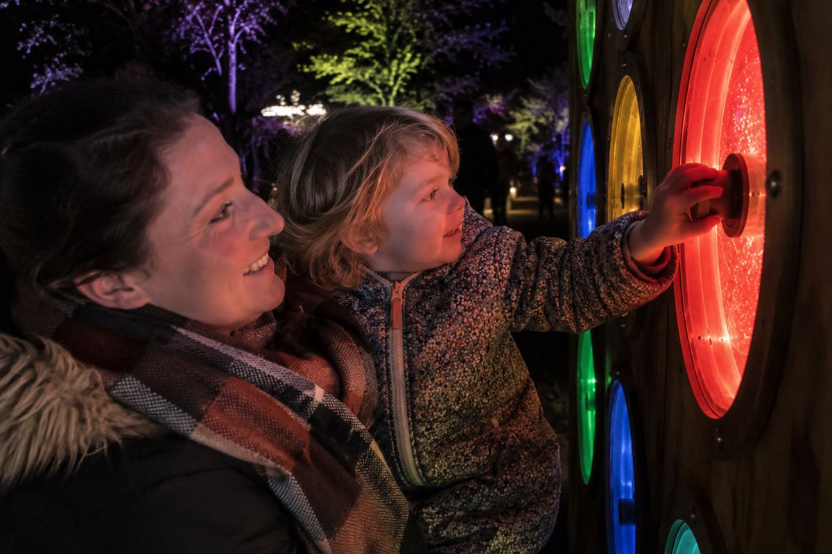 An adult holds a small child up to a panel of glowing coloured discs at night, with colour-lit trees behind them.